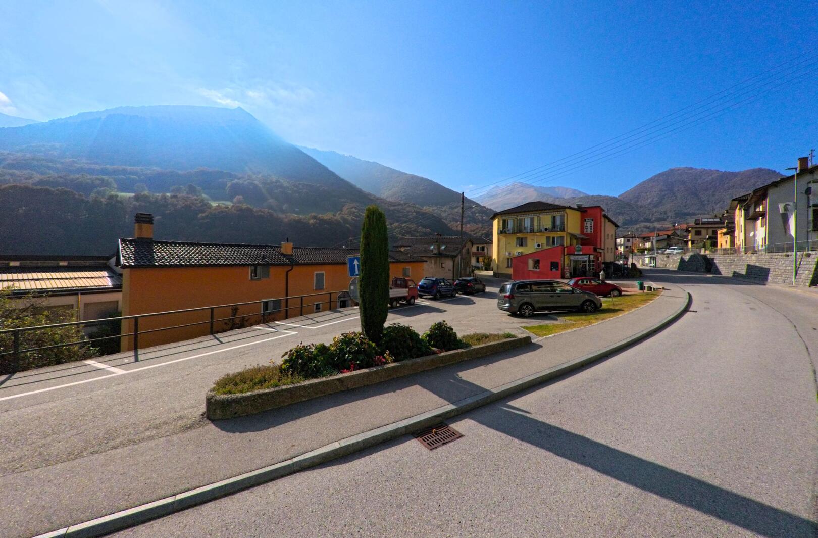 Entlang einer leicht geschwungenen Strasse mit Parkplätzen zeigt sich das Dorfzentrum von Isone (6810, Kanton Tessin) vor bewaldeten Berghängen bei klarem Himmel.