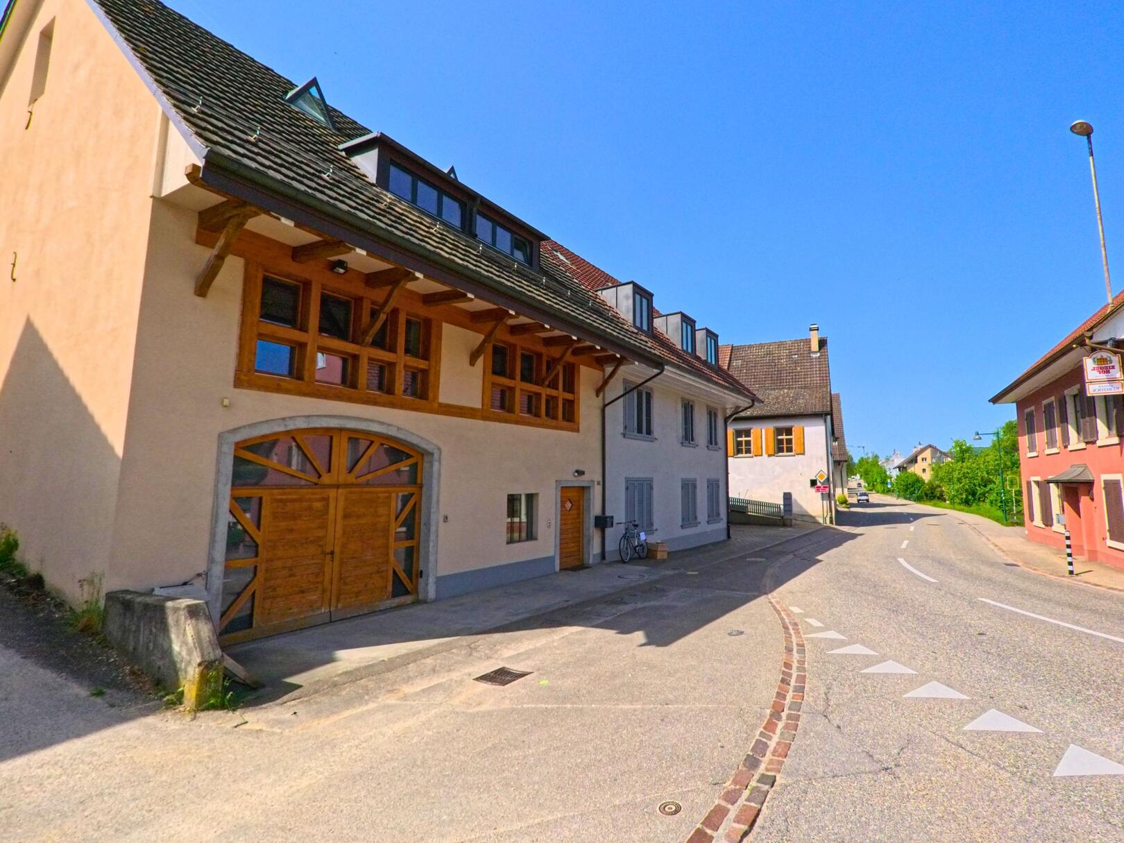 In Itingen (4452, Kanton Baselland) ist eine Ecke mit zwei traditionellen Häusern zu sehen, eines davon hat eine markante Holzstruktur. Die Strasse führt sanft bergauf.