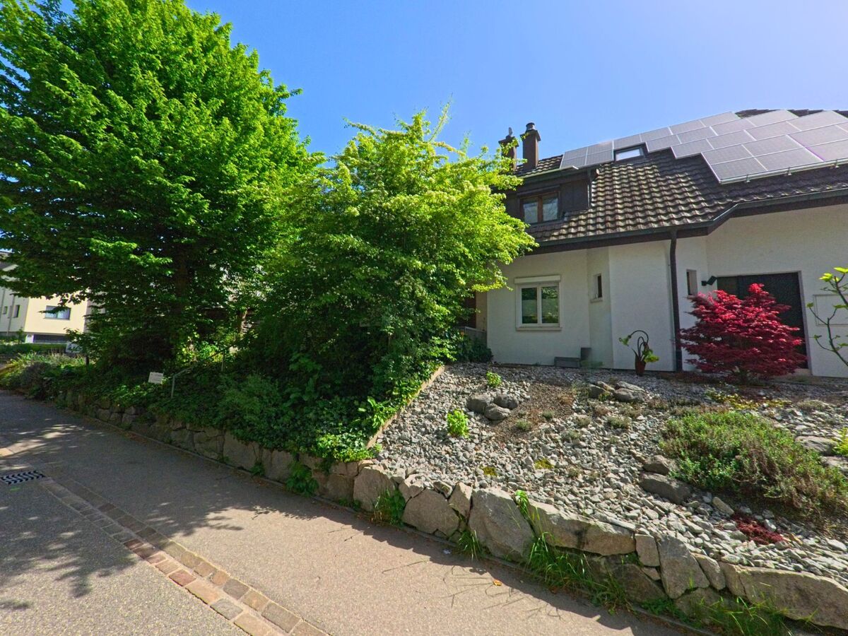 In Itingen (4452, Kanton Baselland) führt eine Straße an einem Haus mit einem bepflanzten Garten und einem Solarpanel auf dem Dach entlang. Bäume und Sträucher säumen den Weg.