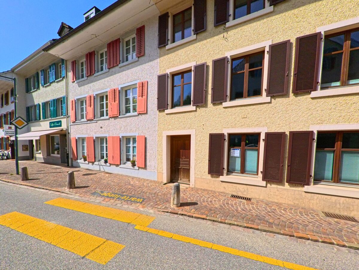 In Itingen (4452, Kanton Baselland) ist ein traditionelles Gebäude mit rosa Fensterläden und einem Geschäft im Erdgeschoss zu sehen. Die Straße verläuft an dem Haus entlang.