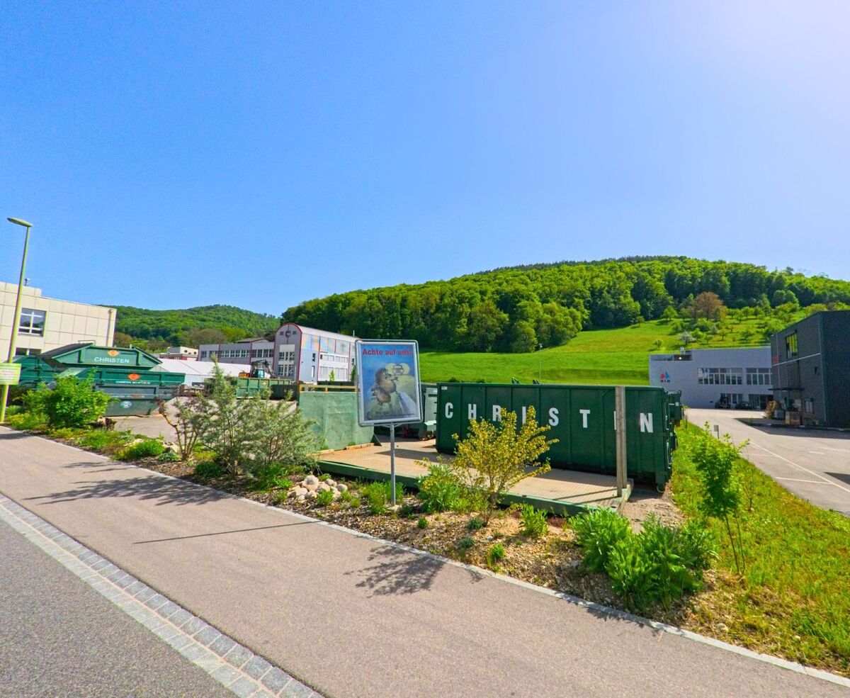In Itingen (4452, Kanton Baselland) ist ein Industriegebäude mit großen Containern im Vordergrund zu sehen. Der Hügel im Hintergrund ergänzt das Bild.