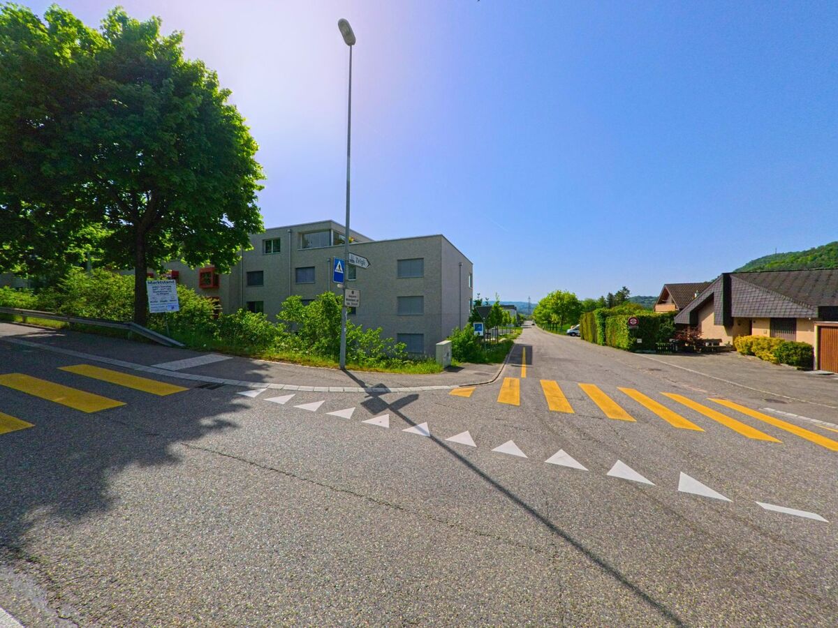 In Itingen (4452, Kanton Baselland) zeigt die Straße eine Kurve, die von Bäumen gesäumt ist, während moderne und traditionelle Häuser im Hintergrund sichtbar sind. Ein Zebrastreifen führt zur nächsten Straße.