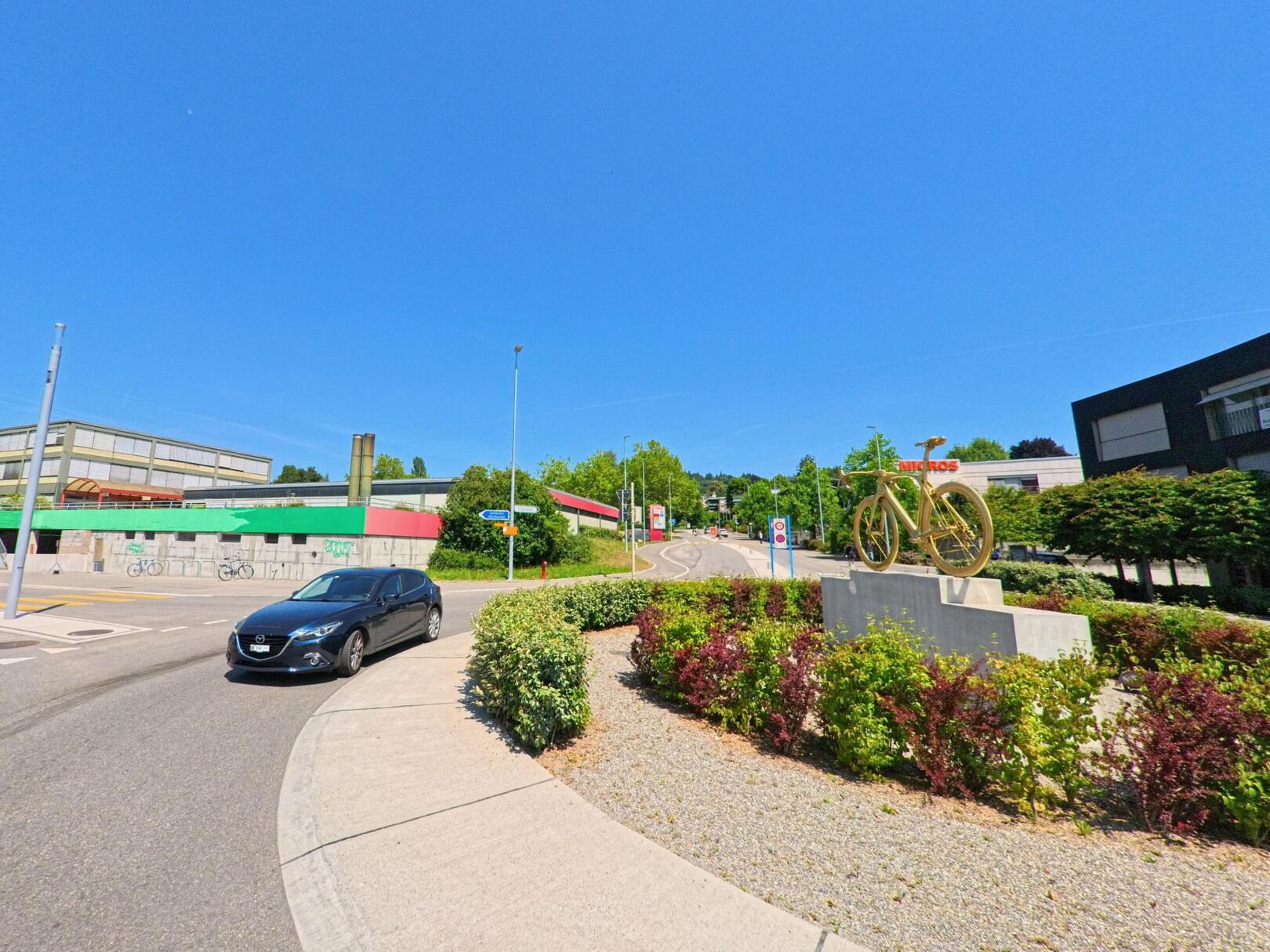 In Ittigen (3063, Kanton Bern) ist ein Kreisverkehr mit einer Fahrradskulptur und modernen Gebäuden zu sehen, während ein Auto langsam durch den Kreisel fährt.