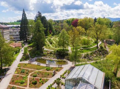 Der Botanische Garten in Freiburg (1701, Kanton Freiburg) zeigt gepflegte Wege, Pflanzenbeete, ein Gewaechshaus und einen kleinen Teich inmitten gruener Baeume.