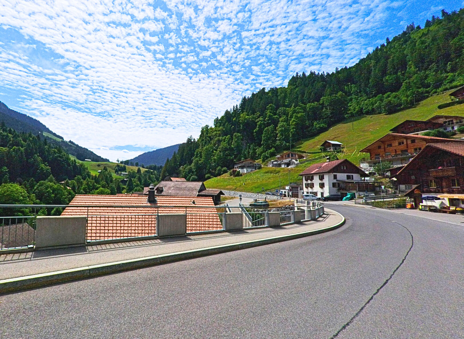 Die kurvige Hauptstrasse von Jaun (1656, Kanton Freiburg) führt vorbei an traditionellen Häusern mit Holzfassaden und bietet einen Blick ins grüne Tal zwischen bewaldeten Berghängen.