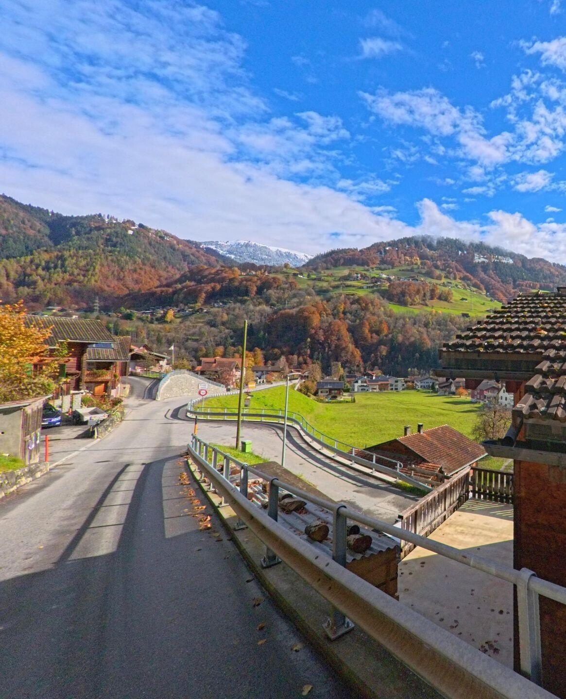 Von einer leicht geschwungenen Dorfstrasse aus zeigt sich in Jenaz (7231, Kanton Graubünden) ein Blick über Wiesen, Wohnhäuser und die herbstlich gefärbten Berghänge.