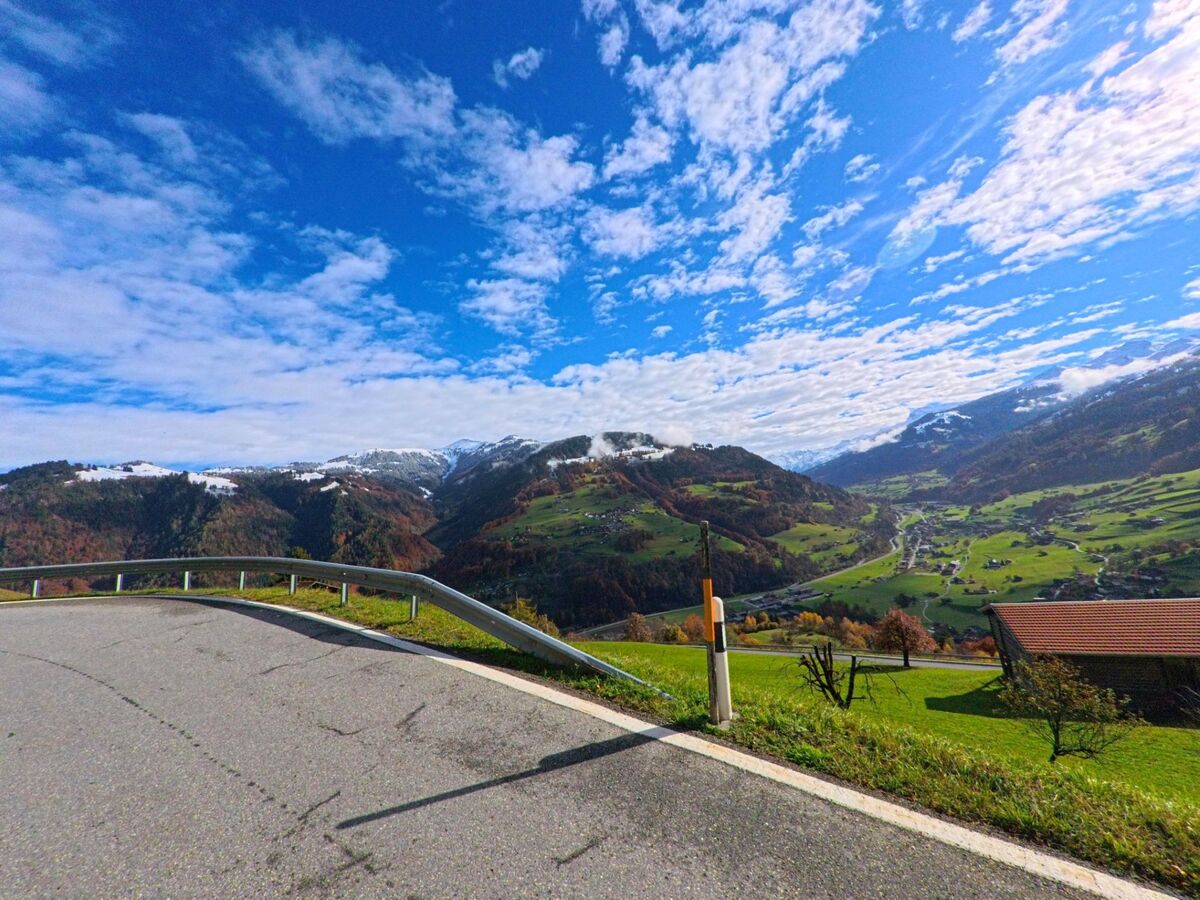 Von einer Kurve an der Hangstrasse eröffnet sich in Jenaz (7231, Kanton Graubünden) ein weiter Blick über das grüne Tal, verstreute Höfe und die herbstlich gefärbten Bergzüge.