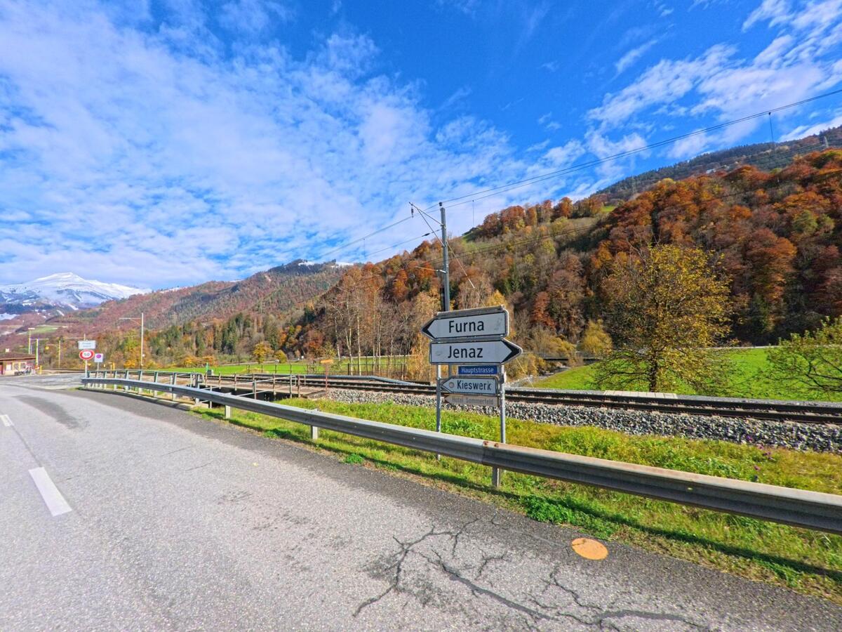 An der Hauptstrasse bei Jenaz (7231, Kanton Graubünden) stehen Wegweiser an einer Bahnlinie, während sich dahinter Wiesen und herbstlich gefärbte Berghänge ausbreiten.
