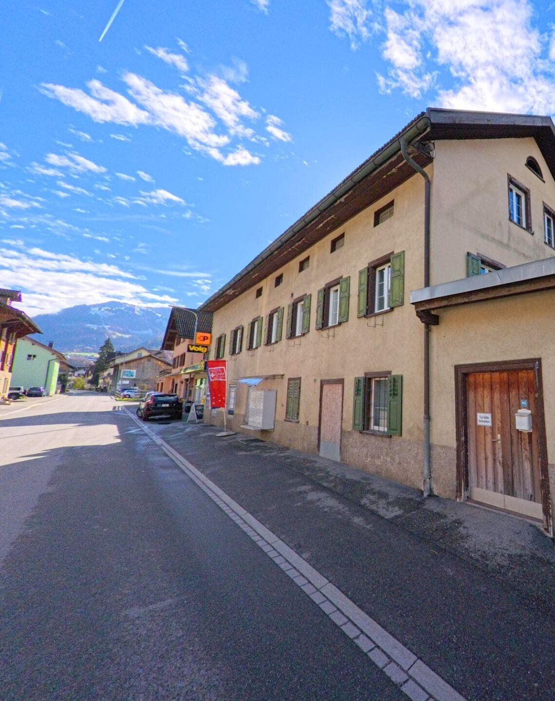 Entlang der Dorfstrasse in Jenaz (7233, Kanton Graubünden) reihen sich ältere Wohn- und Geschäftshäuser mit Fensterläden und Ladenbeschilderung vor der Bergkulisse.