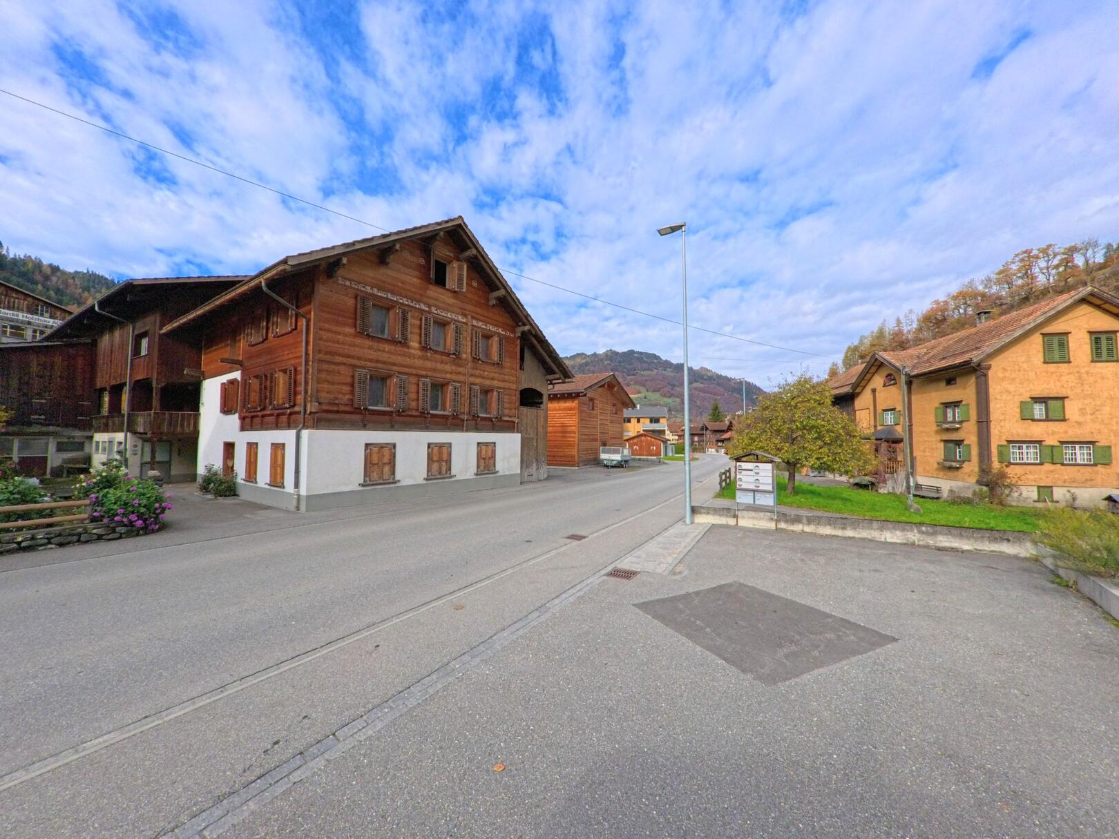 An einer ruhigen Dorfstrasse in Jenaz (7233, Kanton Graubünden) stehen traditionelle Holzhäuser mit Fensterläden neben gepflegten Grünflächen vor der hügeligen Landschaft.