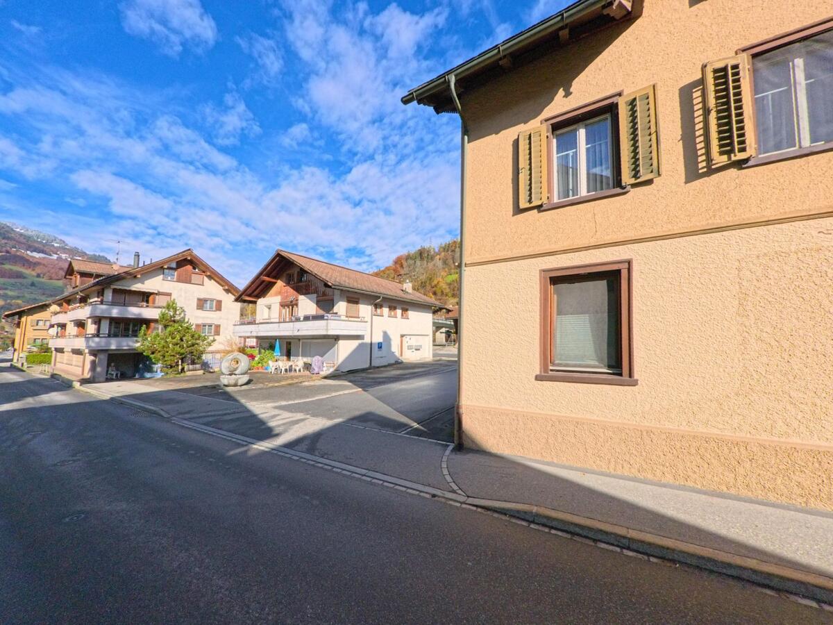 An einer kleinen Kreuzung in Jenaz (7233, Kanton Graubünden) stehen Wohnhäuser mit Balkonen und Fensterläden, eingerahmt von Nebenstrassen und der herbstlichen Hanglage.