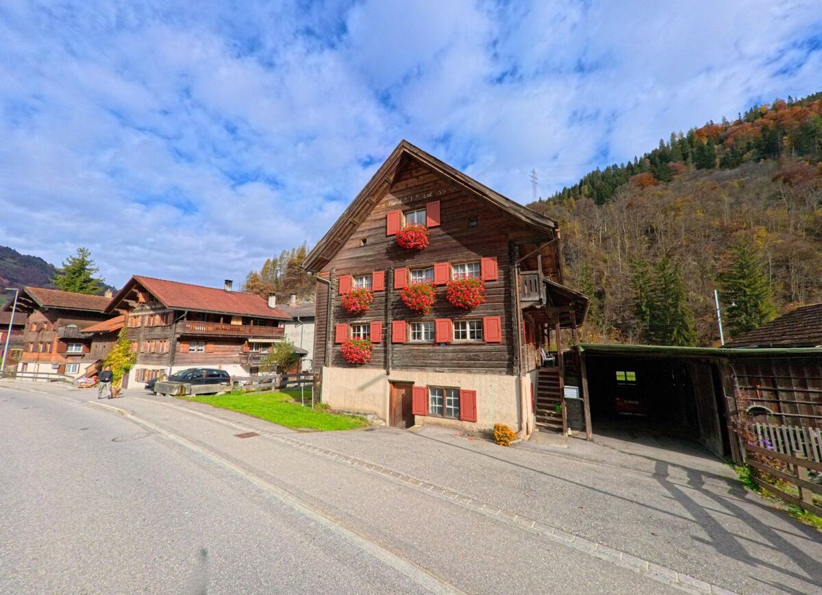 An einer Dorfstrasse in Jenaz (7233, Kanton Graubünden) steht ein traditionelles Holzhaus mit roten Fensterläden und Blumenschmuck vor dem bewaldeten Hang.