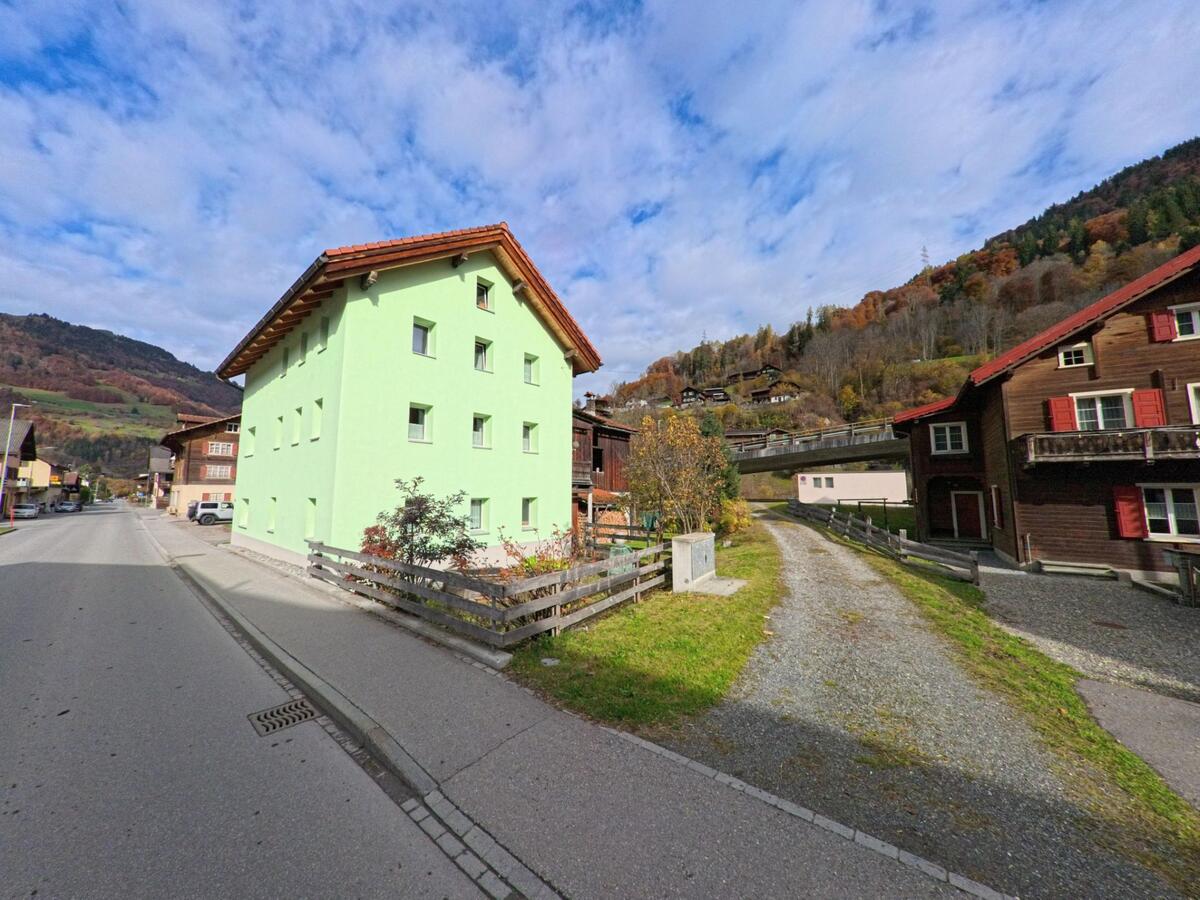 Neben einer Dorfstrasse in Jenaz (7233, Kanton Graubünden) steht ein hell gestrichenes Wohnhaus, während ein schmaler Weg zwischen Holzbauten und Grünflächen ins Quartier führt.