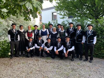 Auf einem Kiesplatz in Davos Glaris (7277, Kanton Graubünden) posiert eine Gruppe in Trachtenkleidung unter grossen Baeumen vor einem hellen Gebaeude.