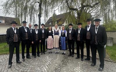 Gruppenfoto des Jodelclubs Schwyzerhüsli Dübendorf in Dübendorf (8600, Kanton Zürich). Die Mitglieder stehen in traditioneller Tracht im Freien vor einer historischen Kulisse mit Bäumen und Gebäuden. Das Bild vermittelt schweizerisches Brauchtum, Gemeinschaft und die Pflege des Jodelgesangs.