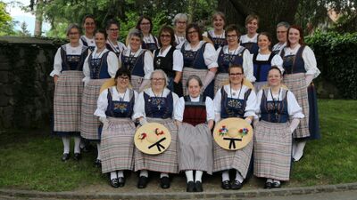 In Kriens (6010, Kanton Luzern) sitzt und steht eine Trachtengruppe in traditionellen Kleidern im Gruenen, waehrend mehrere Mitglieder breite Strohhue­te mit Blumen halten.