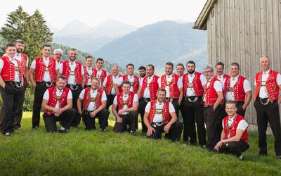 Die Mitglieder des Jodlerklubs Männertreu Nesslau aus Nesslau (9652, Kanton St. Gallen) posieren gemeinsam in traditioneller Tracht mit roten Westen auf einer Wiese vor einer Holzhütte und alpiner Berglandschaft.