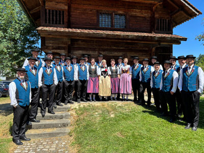 Vor einem traditionellen Holzhaus in Nottwil (6207, Kanton Luzern) stellt sich der Jodlerklub Nottwil in festlicher Tracht zu einem Gruppenfoto im Sonnenschein auf.