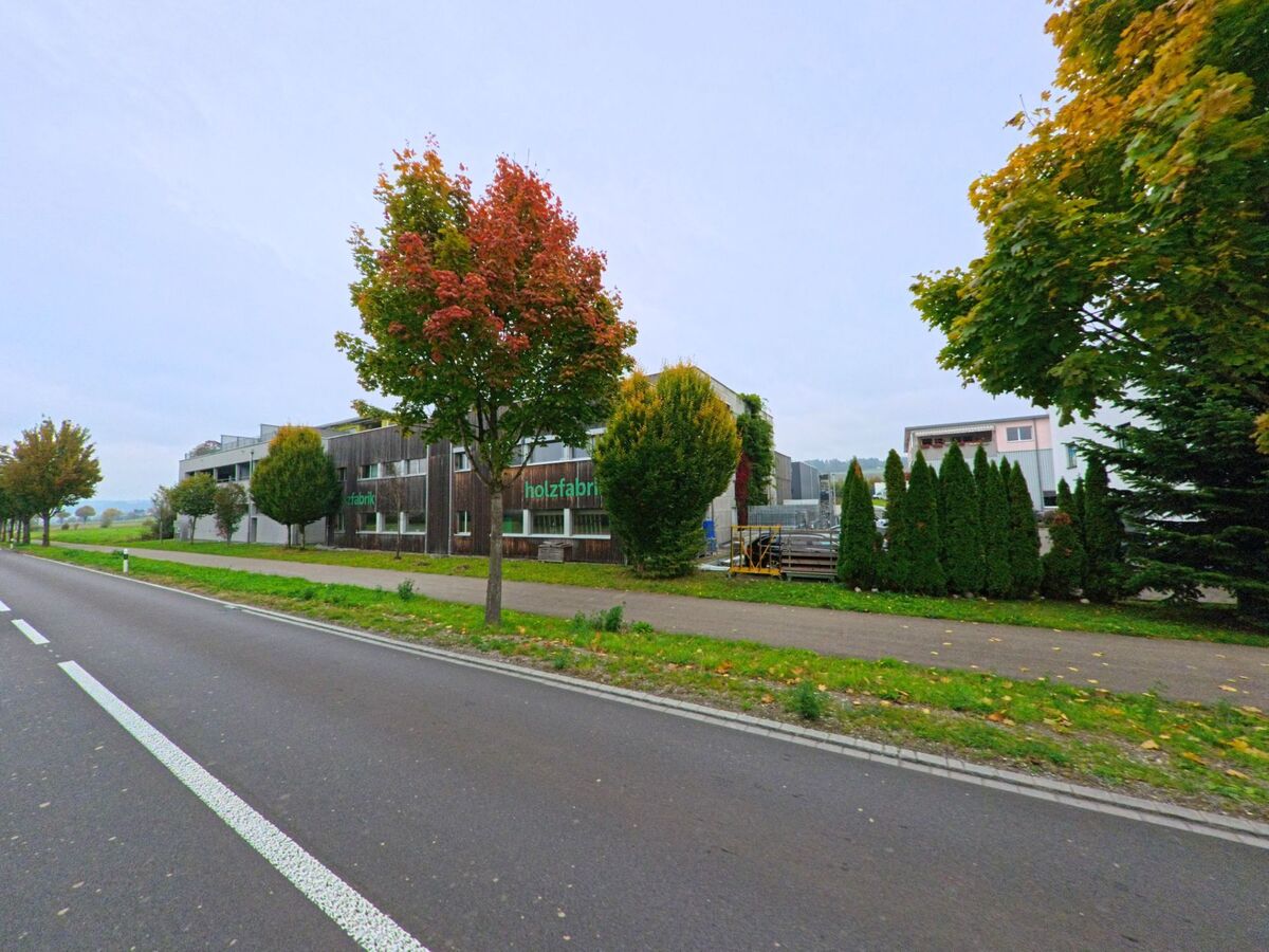 Entlang einer Allee in Jonen (8916, Kanton Aargau) liegt ein modernes Gewerbegebäude mit der Aufschrift „Holzfabrik“, umgeben von herbstlich gefärbten Bäumen.