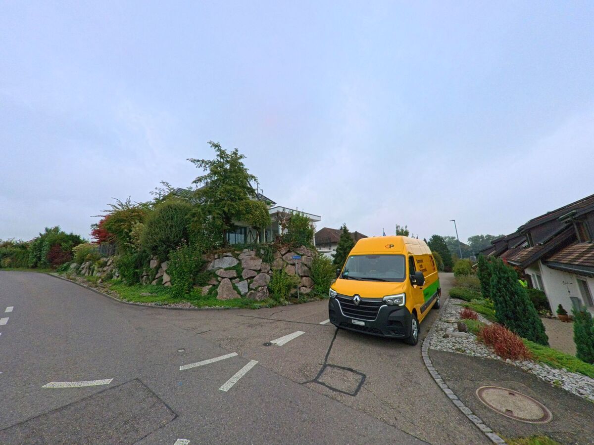 Ein gelber Lieferwagen der Post steht in einer Wohnstrasse in Jonen (8916, Kanton Aargau) vor einem Haus mit bepflanzter Natursteinmauer.