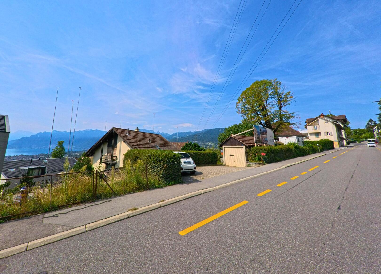 Entlang einer Strasse in Jongny (1805, Kanton Waadt) stehen Einfamilienhaeuser mit Blick auf den Genfersee und die Berge im Hintergrund.