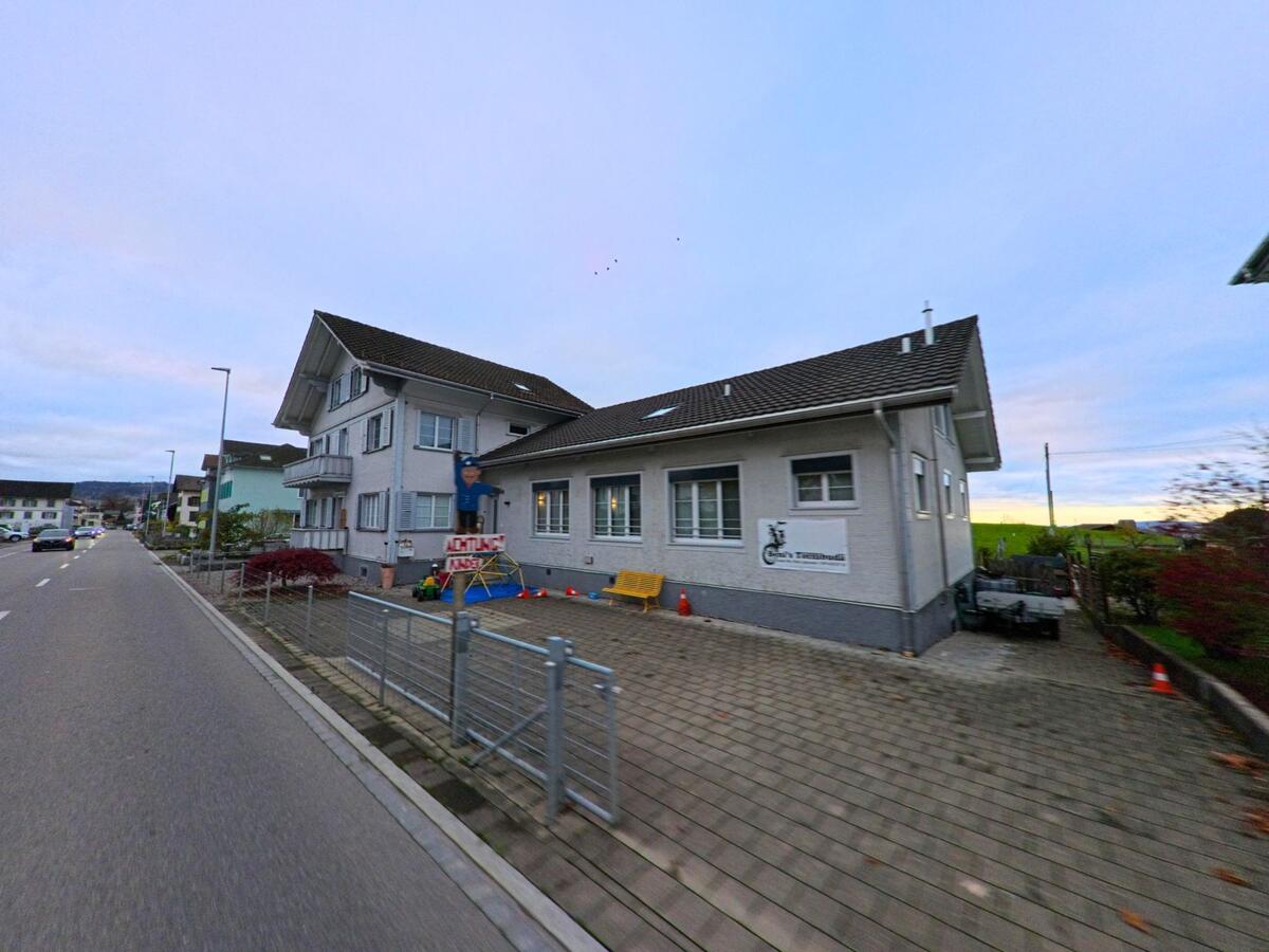 An einer Strasse in Jonschwil (9243, Kanton St. Gallen) steht ein helles Gebaeude mit Spielbereich im Vorplatz und Blick auf die offenen Wiesen im Hintergrund.