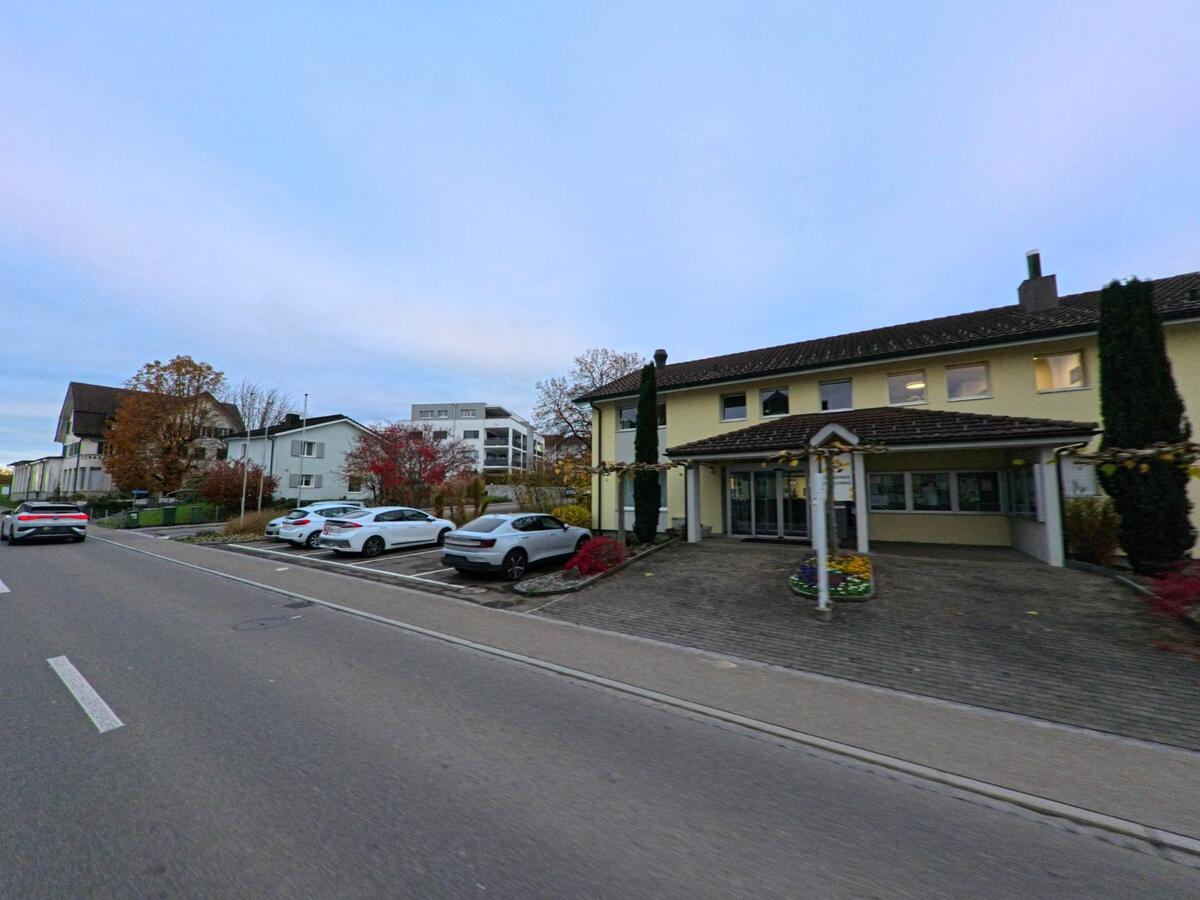 Ein hellgelbes Gebaeude in Jonschwil (9243, Kanton St. Gallen) liegt an der Strasse, davor mehrere Parkplaetze und herbstlich bepflanzte Beete mit bunten Blumen.