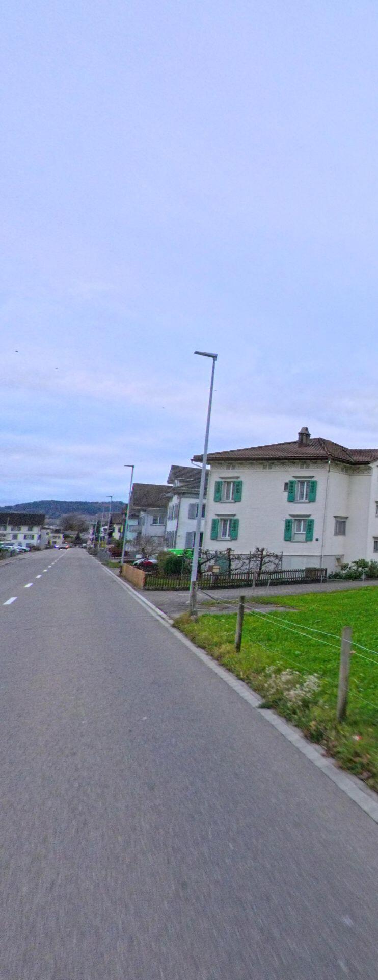 Eine lange, leicht geschwungene Strasse in Jonschwil (9243, Kanton St. Gallen) verlaeuft an Wohnhaeusern mit gruenen Fensterlaeden vorbei und fuehrt in Richtung der Huegellandschaft.