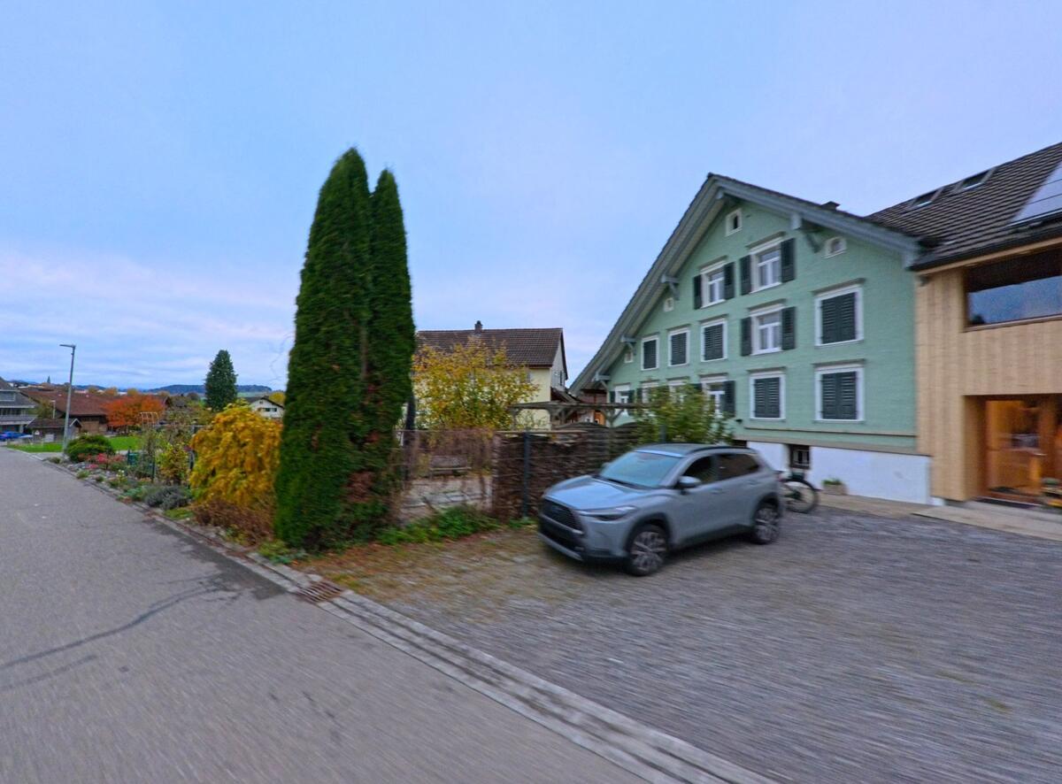 Ein mintfarbenes Wohnhaus in Jonschwil (9243, Kanton St. Gallen) steht an einer ruhigen Strasse, daneben ein geparkter Wagen und ein Garten mit herbstlicher Bepflanzung.