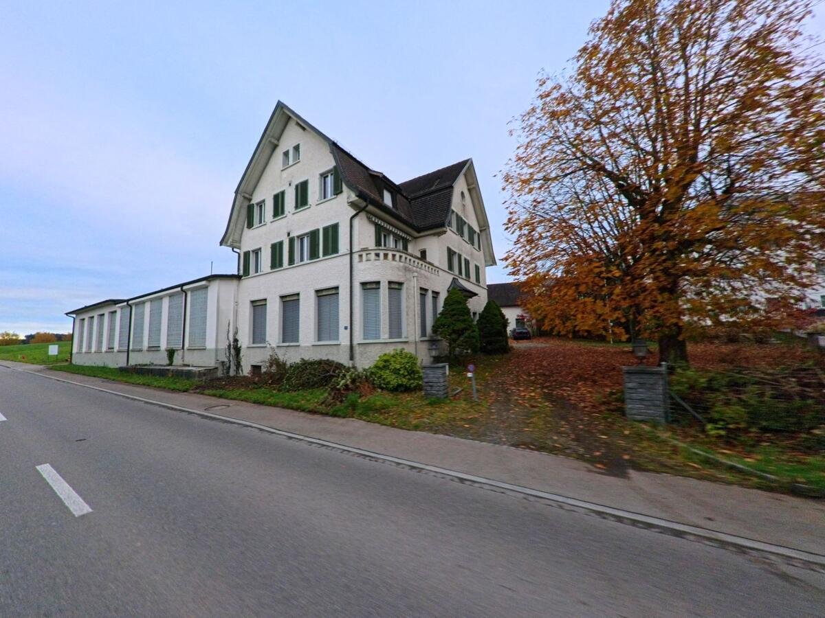 Ein grosses weisses Gebaeude in Jonschwil (9243, Kanton St. Gallen) steht an einer Strasse, daneben eine von Herbstlaub bedeckte Einfahrt mit einem grossen Baum.