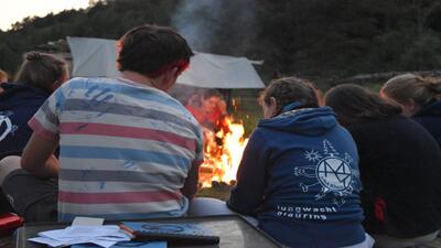 In Bülach (8180, Kanton Luzern) sitzen mehrere Kinder und Betreuer um ein prasselndes Lagerfeuer, während sie die flimmernden Flammen beobachten und im Hintergrund das Zelt sichtbar ist.