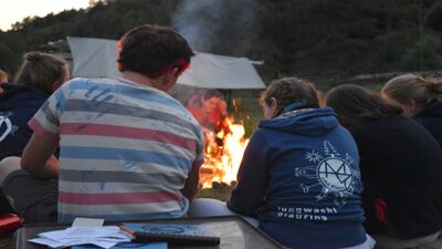 In Bülach (8180, Kanton Luzern) sitzen mehrere Kinder und Betreuer um ein prasselndes Lagerfeuer, während sie die flimmernden Flammen beobachten und im Hintergrund das Zelt sichtbar ist.