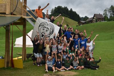 In Neuheim (6345, Kanton Zug) sitzt und steht die Jubla Neuheim als grosse Kinder- und Jugendgruppe in einheitlichen dunkelblauen Shirts in einer Sporthalle.