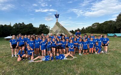 Die Kinder und Jugendlichen der Jubla Sarmenstorf posieren in blauen Vereins-T-Shirts vor einem grossen Zeltlager auf einer Wiese in Sarmenstorf (5614, Kanton Aargau).