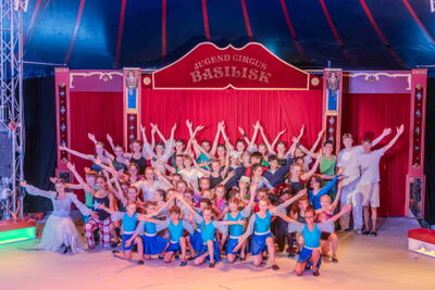 Auf einer beleuchteten Zirkusbuehne stehen in Basel (4052, Kanton Basel-Stadt) zahlreiche Kinder und Jugendliche des Jugend Circus gemeinsam in farbigen Kostuemen vor dem roten Vorhang.