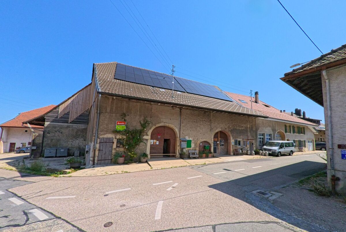 In Juriens (1326, Kanton Waadt) steht ein traditionelles Steingebäude mit grossen Rundbogenportalen und Solarpaneelen auf dem Dach an einer Kreuzung.