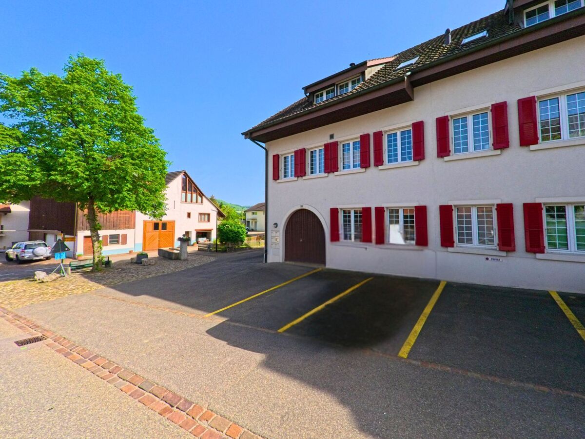 In Kaenerkinden (4447, Kanton Baselland) befindet sich ein Wohnhaus mit roten Fensterlaeden an einem gepflasterten Hof mit markierten Parkplaetzen und umgebenden Nebengebaeuden.