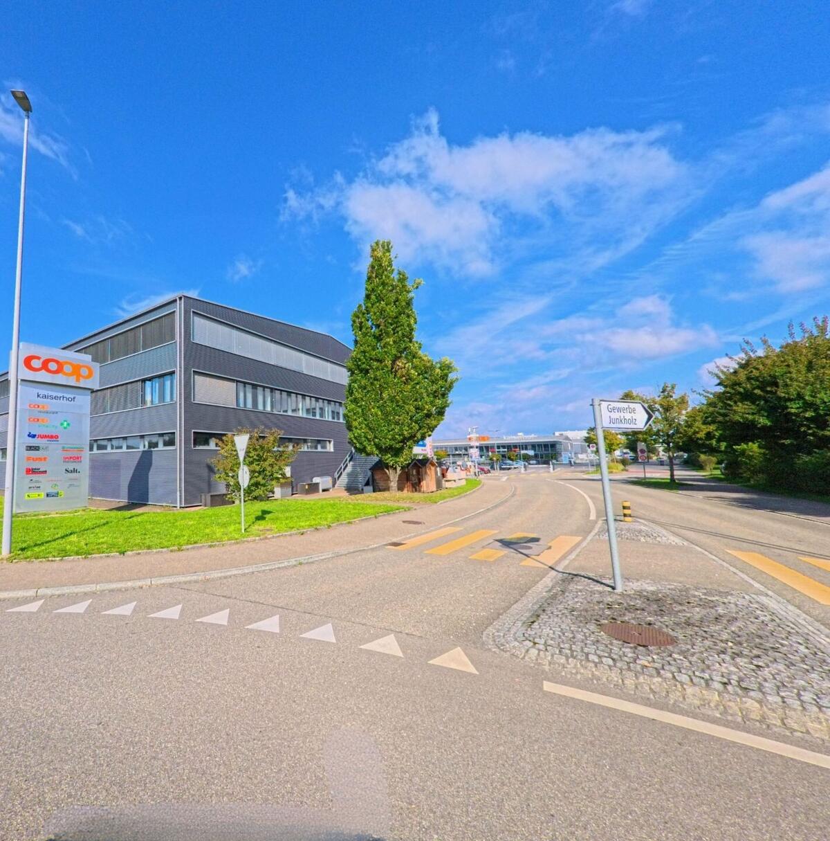 An der Strasse Gewerbe Junkholz in Kaiseraugst (4303, Kanton Aargau) steht ein modernes Gebäude mit Coop-Schild und mehreren Gewerbebetrieben unter blauem Himmel.
