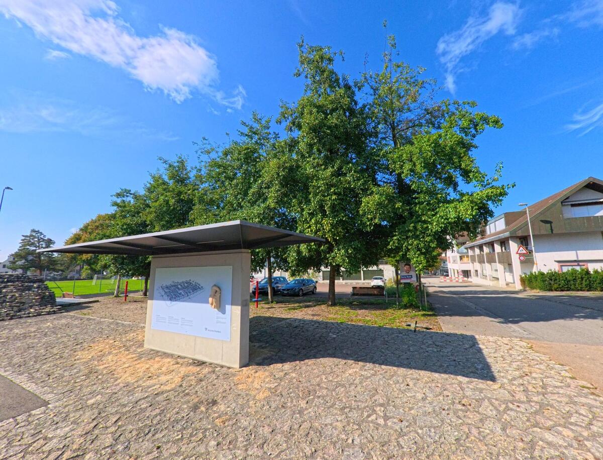Eine Informationstafel mit Dach steht in Kaiseraugst (4303, Kanton Aargau) auf einem gepflasterten Platz, umgeben von Bäumen und Wohnhäusern, bei sonnigem Wetter.