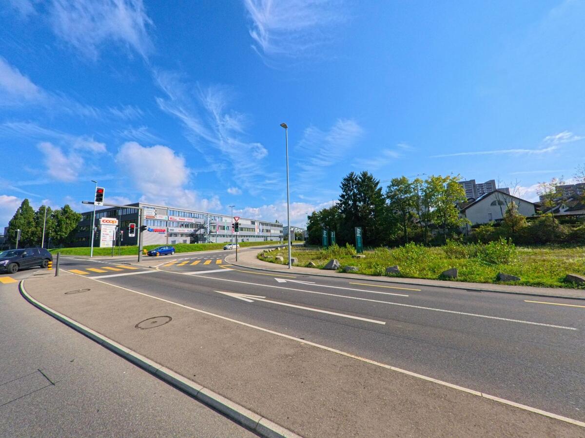 An einer Kreuzung in Kaiseraugst (4303, Kanton Aargau) führt die Strasse an einem modernen Einkaufszentrum mit Coop-Filiale vorbei, während Autos an der Ampel warten.