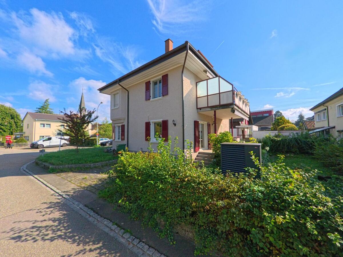 Ein helles Wohnhaus mit roten Fensterläden und Balkon steht in Kaiseraugst (4303, Kanton Aargau) an einer kleinen Strasse, umgeben von Gärten und weiteren Häusern.