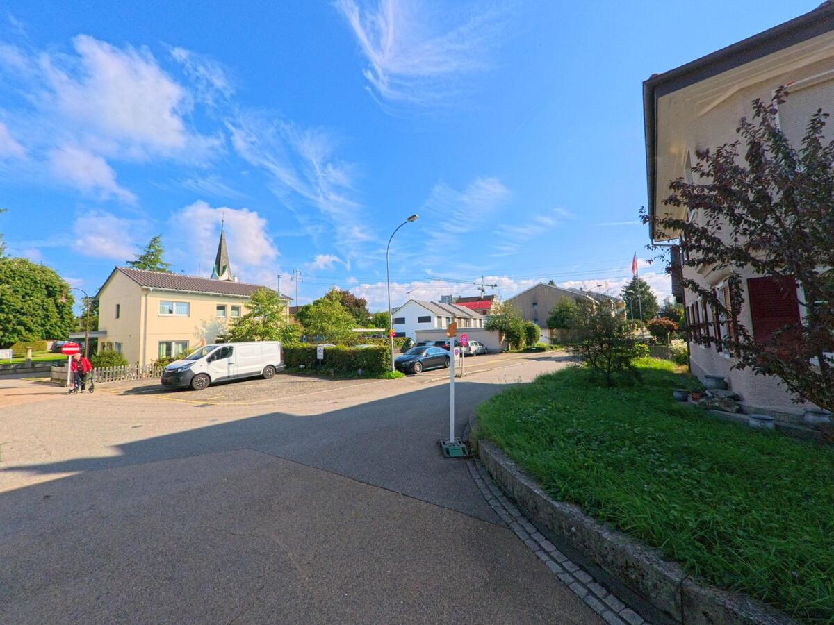 Eine kleine Wohnstrasse in Kaiseraugst (4303, Kanton Aargau) mit Einfamilienhäusern, geparkten Fahrzeugen und Blick auf den Kirchturm unter blauem Himmel.