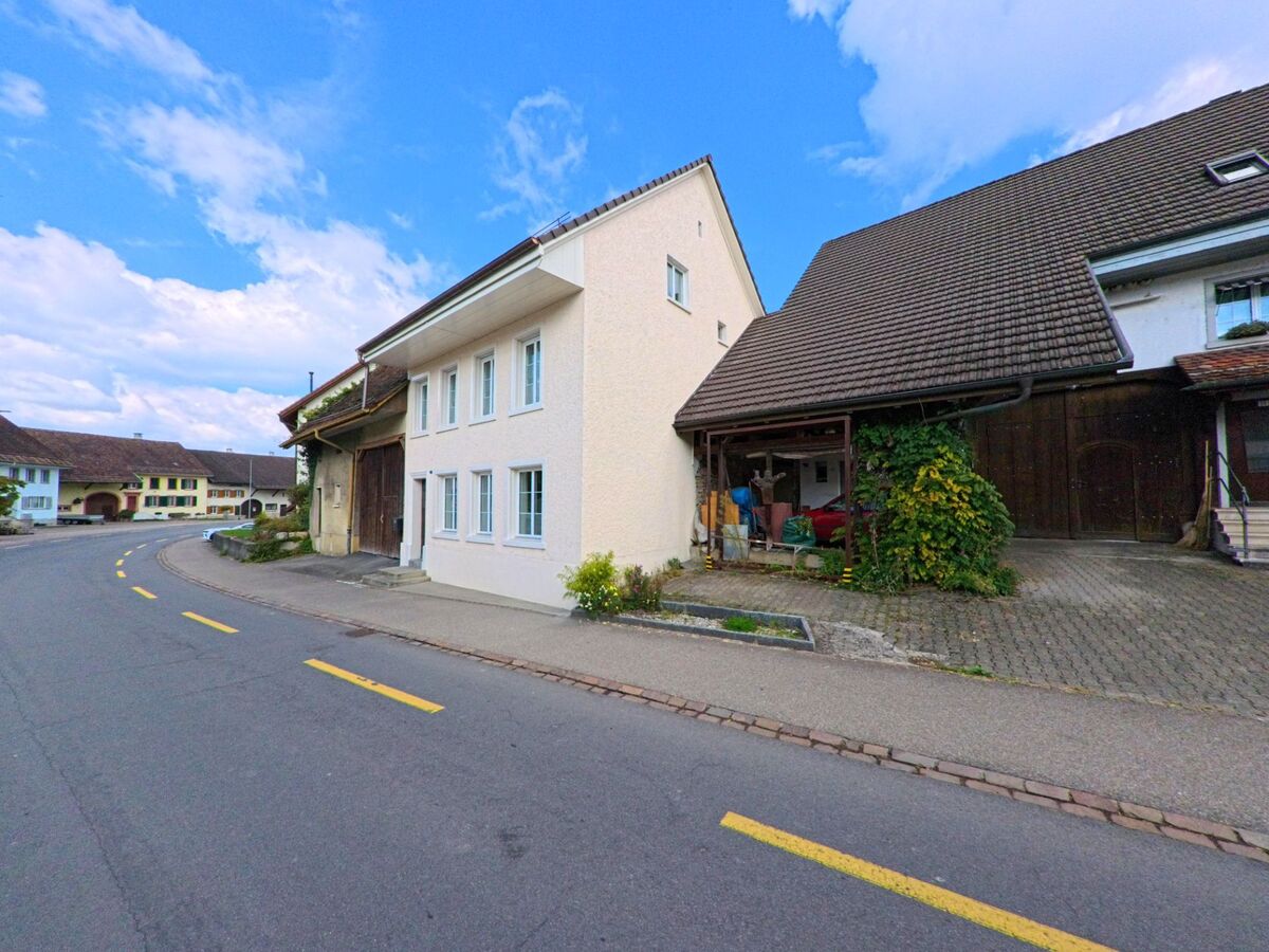 Ein Bauernhaus mit angebauter Scheune steht an einer sanft gebogenen Strasse in Kaisten (5082, Kanton Aargau), im Hintergrund sind weitere traditionelle Gebäude zu sehen.