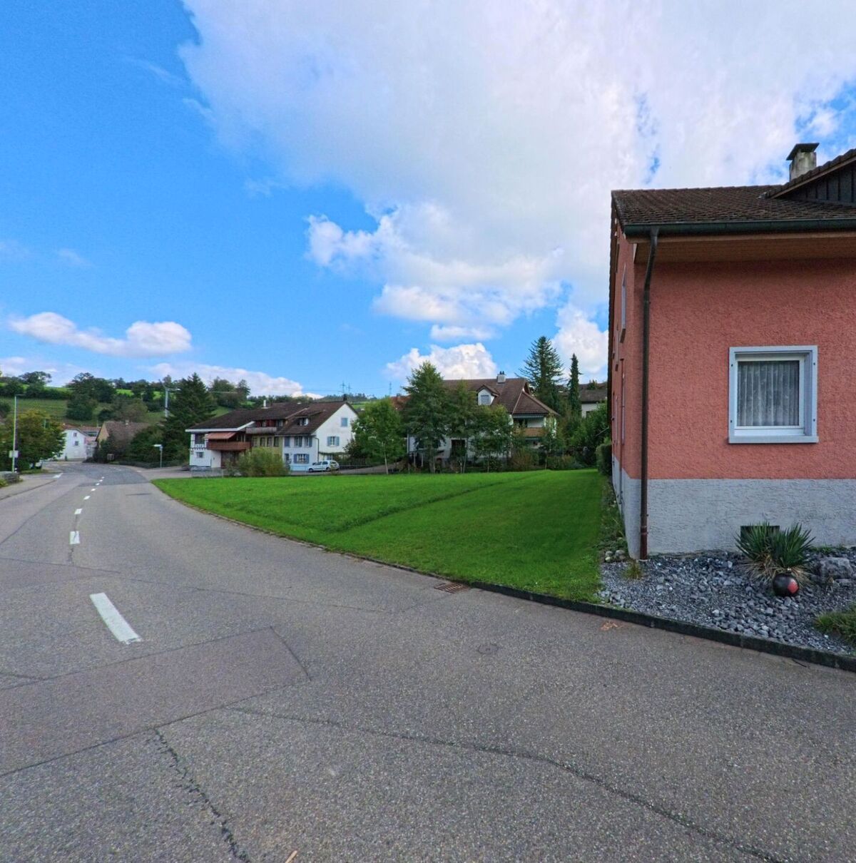 Ein rosafarbenes Wohnhaus steht an einer sanft geschwungenen Strasse in Kaisten (5082, Kanton Aargau), im Hintergrund sind weitere Häuser und grüne Hügel zu sehen.