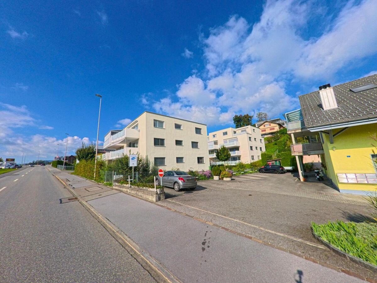 Am Rand von Kaltbrunn (8722, Kanton St. Gallen) stehen moderne Mehrfamilienhaeuser oberhalb einer breiten Strasse mit Gehweg und vereinzeltem Verkehr.