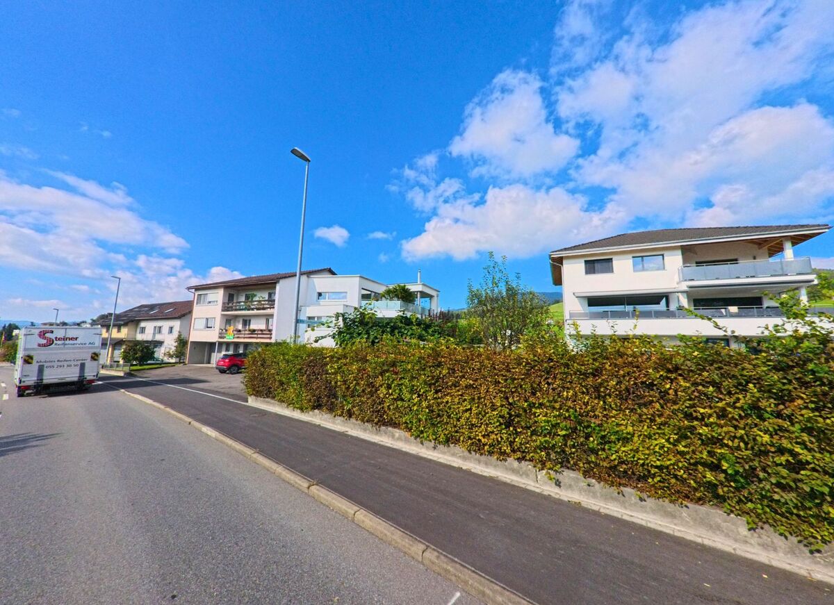 Mehrfamilienhaeuser in Kaltbrunn (8722, Kanton St. Gallen) reihen sich entlang einer breiten Strasse, getrennt durch eine dichte Hecke und einen Gehweg.