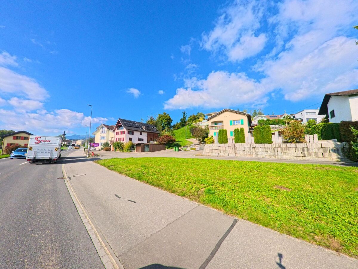 Entlang einer leicht ansteigenden Strasse in Kaltbrunn (8722, Kanton St. Gallen) stehen verschiedene Wohnhaeuser oberhalb eines breiten Gruenstreifens.