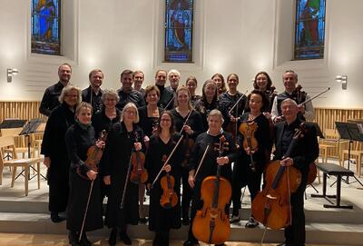 Mitglieder des Kammerorchesters Regensdorf stehen mit Violinen, Bratschen und Celli in einer Kirche für ein Gruppenfoto bereit. Das Ensemble präsentiert sich in Konzertkleidung vor den Kirchenfenstern und dem Aufführungsraum.