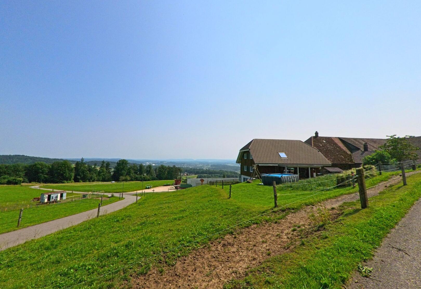 Auf einem sanften Huegel in Kammersrohr (4535, Kanton Solothurn) stehen Bauernhaeuser mit weitem Blick ueber gruenes Weideland bis zum Horizont.