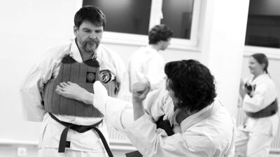 In Aarau (5000, Kanton Aargau) ueben zwei Personen in Weissguerteln und Schutzausruestung einen kraftvollen Tritt im Trainingsraum, waehrend weitere Teilnehmende im Hintergrund trainieren.